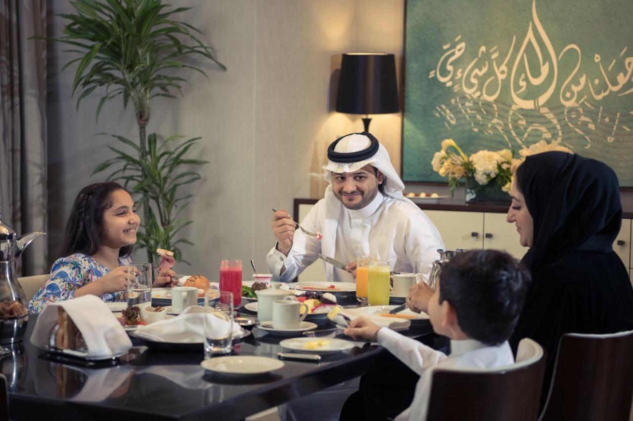 Jabal Omar Hyatt Regency Makkah Hotel Mecca Exterior foto The photo depicts a cheerful family gathered around a dining table for a meal. They appear to be enjoying breakfast or a brunch. The table is set with various dishes and drinks, including juices and plates of food. 

A man wearing traditional attire 