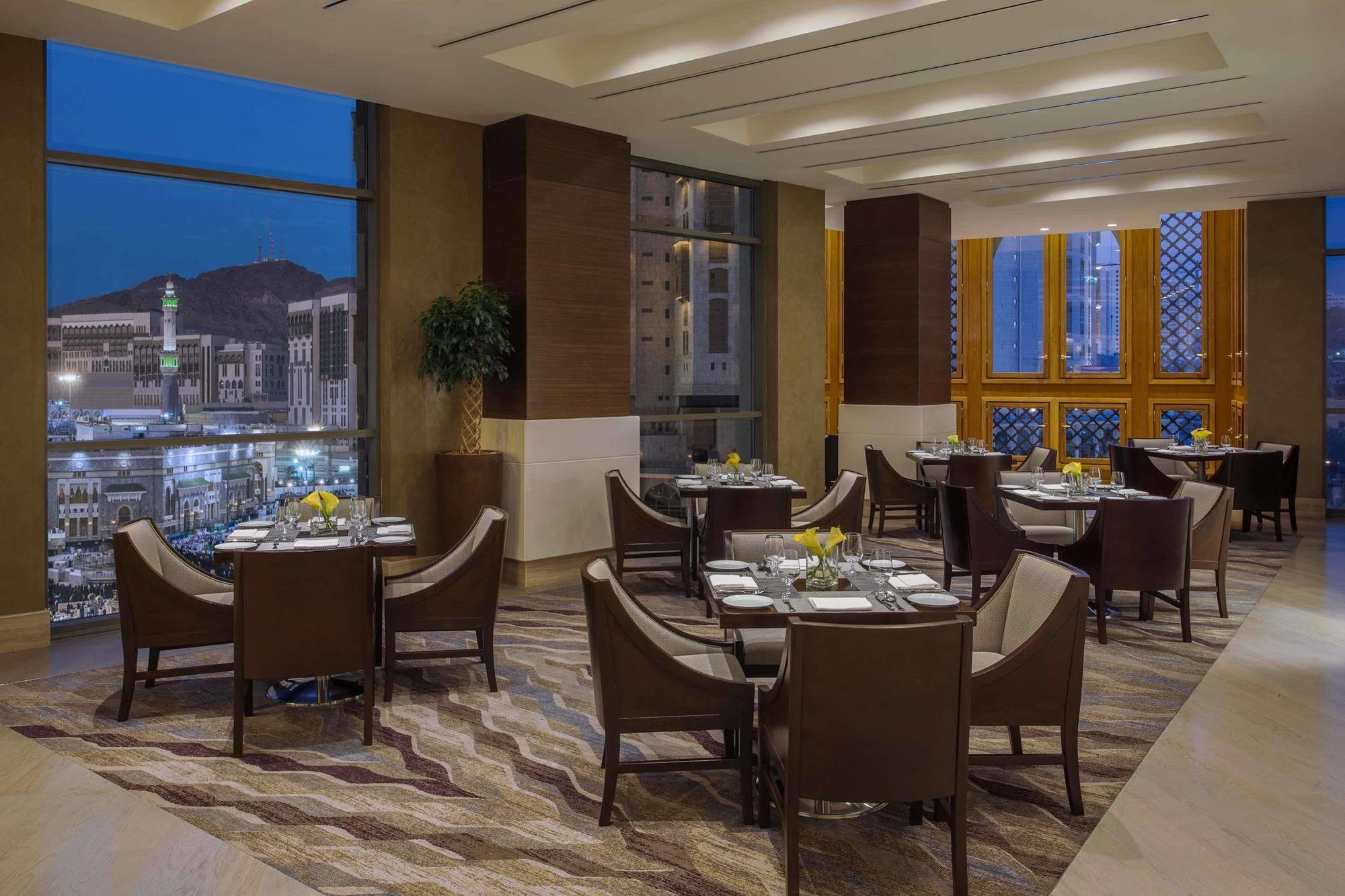 Jabal Omar Hyatt Regency Makkah Hotel Mecca Exterior foto The photo shows a sophisticated restaurant interior, featuring elegant round tables with high-backed chairs arranged neatly. The space is well-lit with large windows that provide a view of the city skyline at dusk, with mountains in the background. T