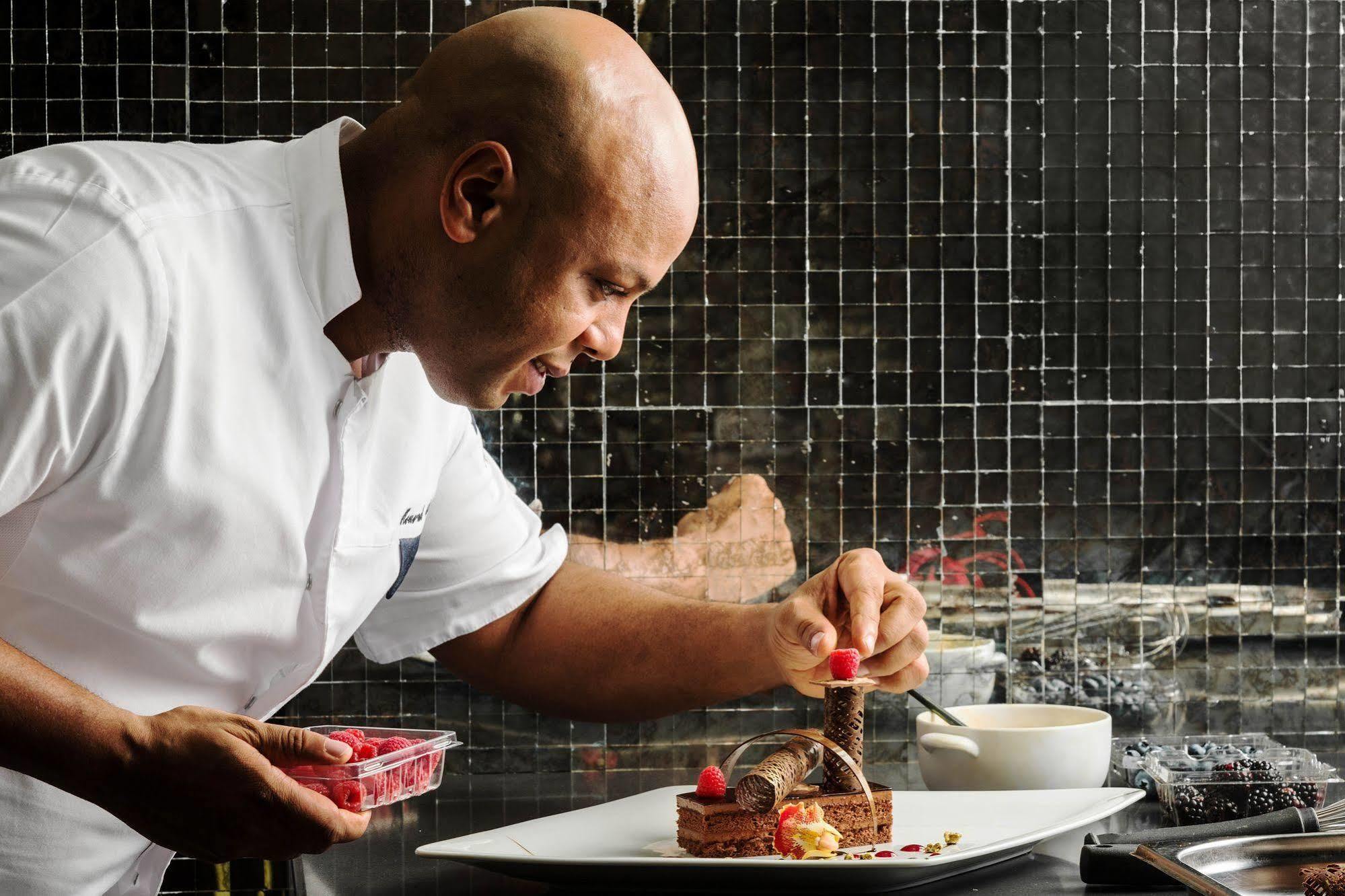 Jabal Omar Hyatt Regency Makkah Hotel Mecca Exterior foto The photo depicts a chef, who is bald and wearing a white chef's coat, carefully plating a dessert. He is focused on adding a finishing touch, possibly a raspberry, to the dessert on a large white plate. The background features a tiled wall, and ther