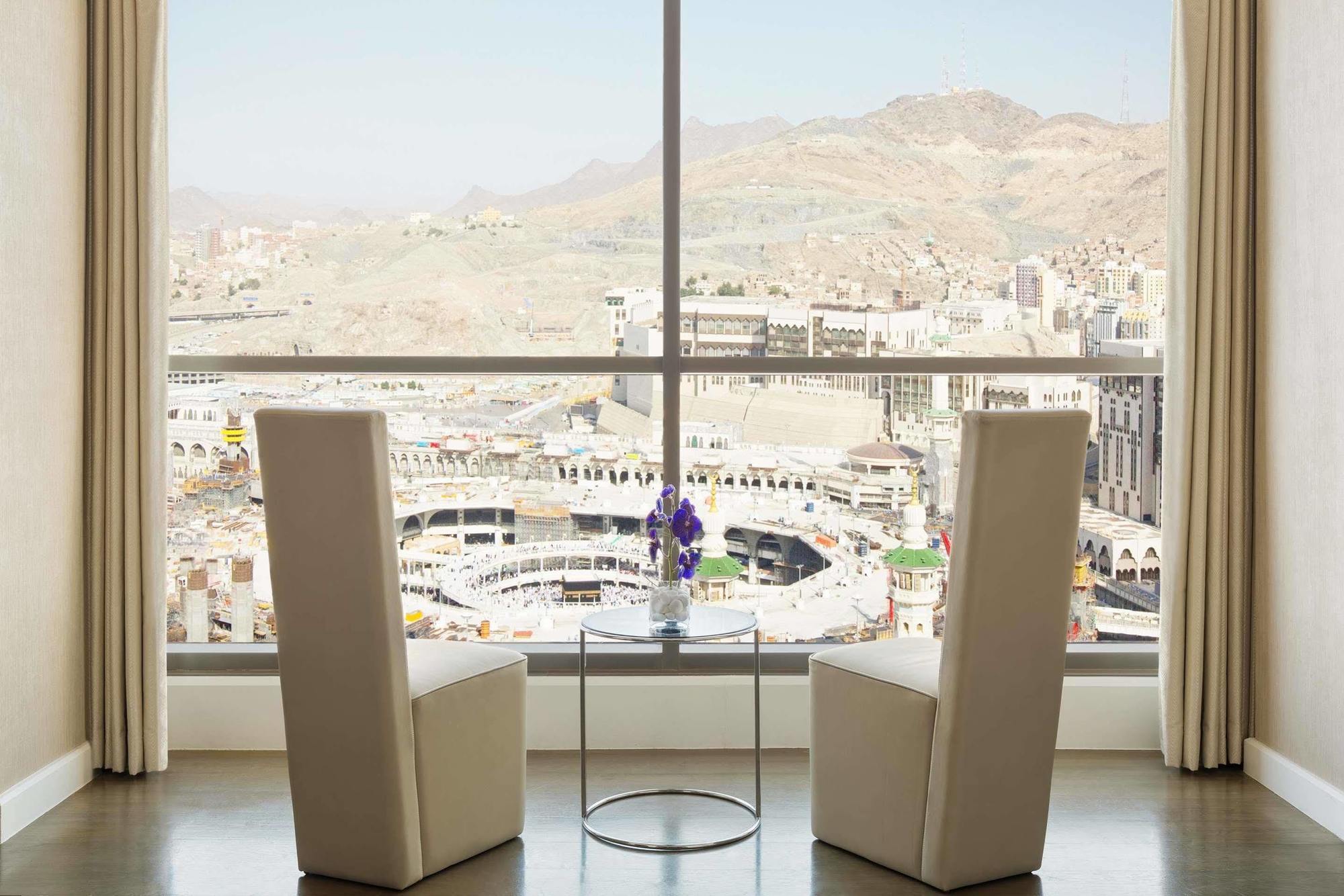 Jabal Omar Hyatt Regency Makkah Hotel Mecca Exterior foto The photo shows a modern indoor setting with two tall, white chairs positioned facing a large window. Through the window, there is a panoramic view of a cityscape that includes mountainous terrain and buildings, suggesting it may be a notable locatio