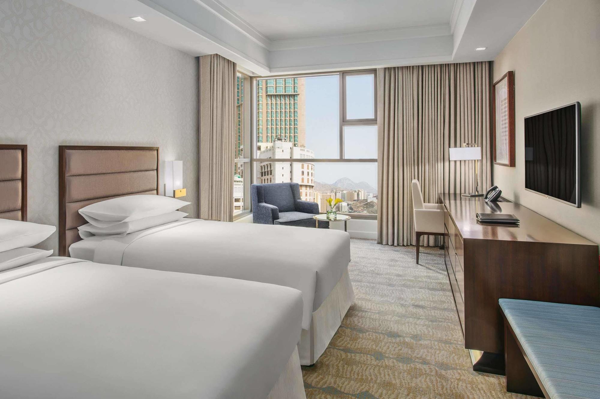 Jabal Omar Hyatt Regency Makkah Hotel Mecca Exterior foto The photo shows a modern hotel room featuring two single beds with white bedding. There's a spacious window allowing natural light to fill the room, which is adorned with light-colored curtains. In one corner, there's a comfortable armchair next to a