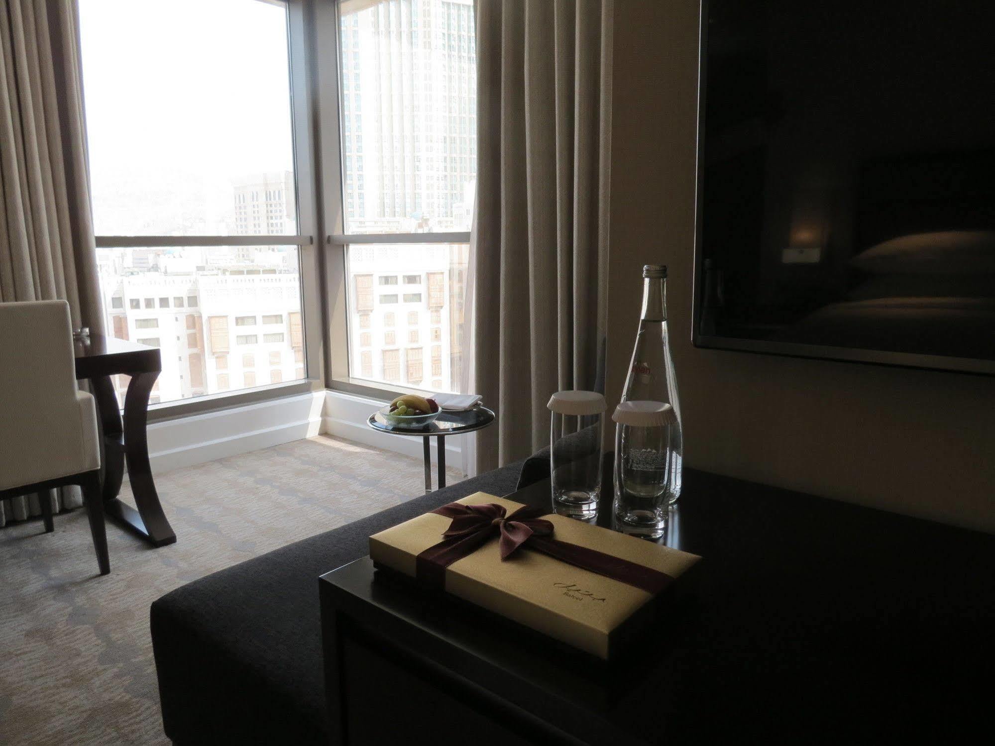 Jabal Omar Hyatt Regency Makkah Hotel Mecca Exterior foto The photo shows a modern hotel room with large windows letting in natural light. In the foreground, there is a table with a decorative box tied with a ribbon, two clear glass water bottles, and a couple of glasses. In the background, there's a comfor