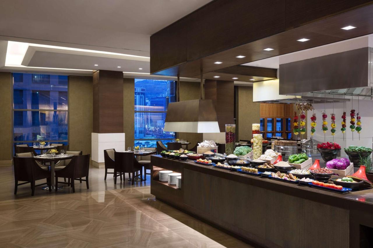 Jabal Omar Hyatt Regency Makkah Hotel Mecca Exterior foto The photo shows a dining area, likely within a restaurant or buffet setting. On the right side, there is a long food display featuring various dishes, likely consisting of fruits, vegetables, and other culinary items. The area is well-lit, with moder